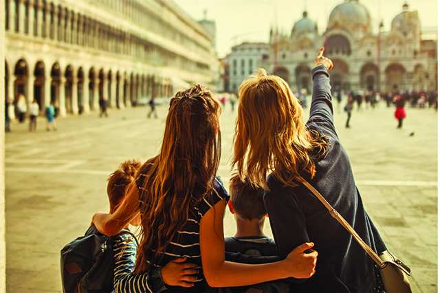 Mother and three kids visiting Venice, Italy. They are standing in Piazza San Marco. Mother is showing them the Basilica di San Marco and the Campanile di San Marco. Sunny day in Venice, Italy.