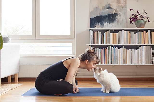 woman kisses a cats head while both seated on a yoga mat