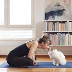 woman kisses a cats head while both seated on a yoga mat